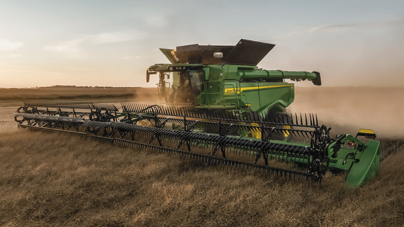 John Deere combine harvesting grain in a dusty field at sunset using a wide draper header. John Deere combine harvesting grain in a dusty field at sunset using a wide draper header.