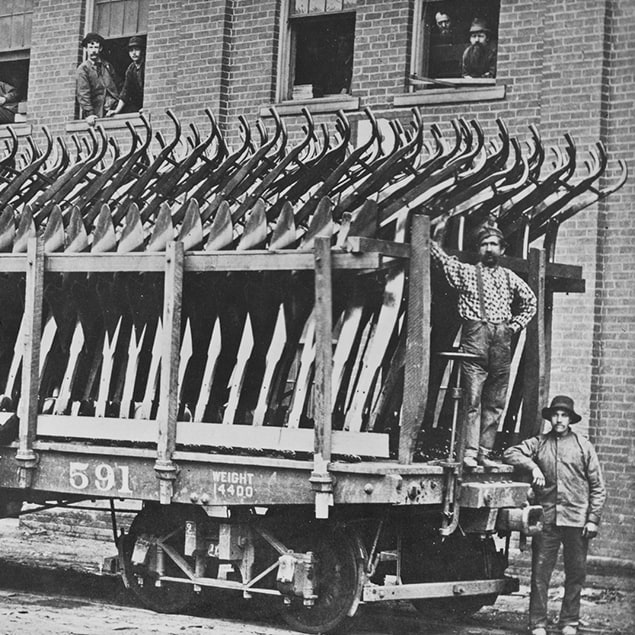 出荷準備の整った鋼鉄製プラウを積んだ Deere & Co の列車の横に立つ 3 人の男性と、背後の建物の窓から外を見る工場従業員たちの 1882 年の写真