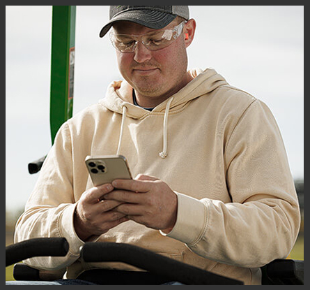 A person sitting on a zero-turn mower with safety glasses on looking at a phone.