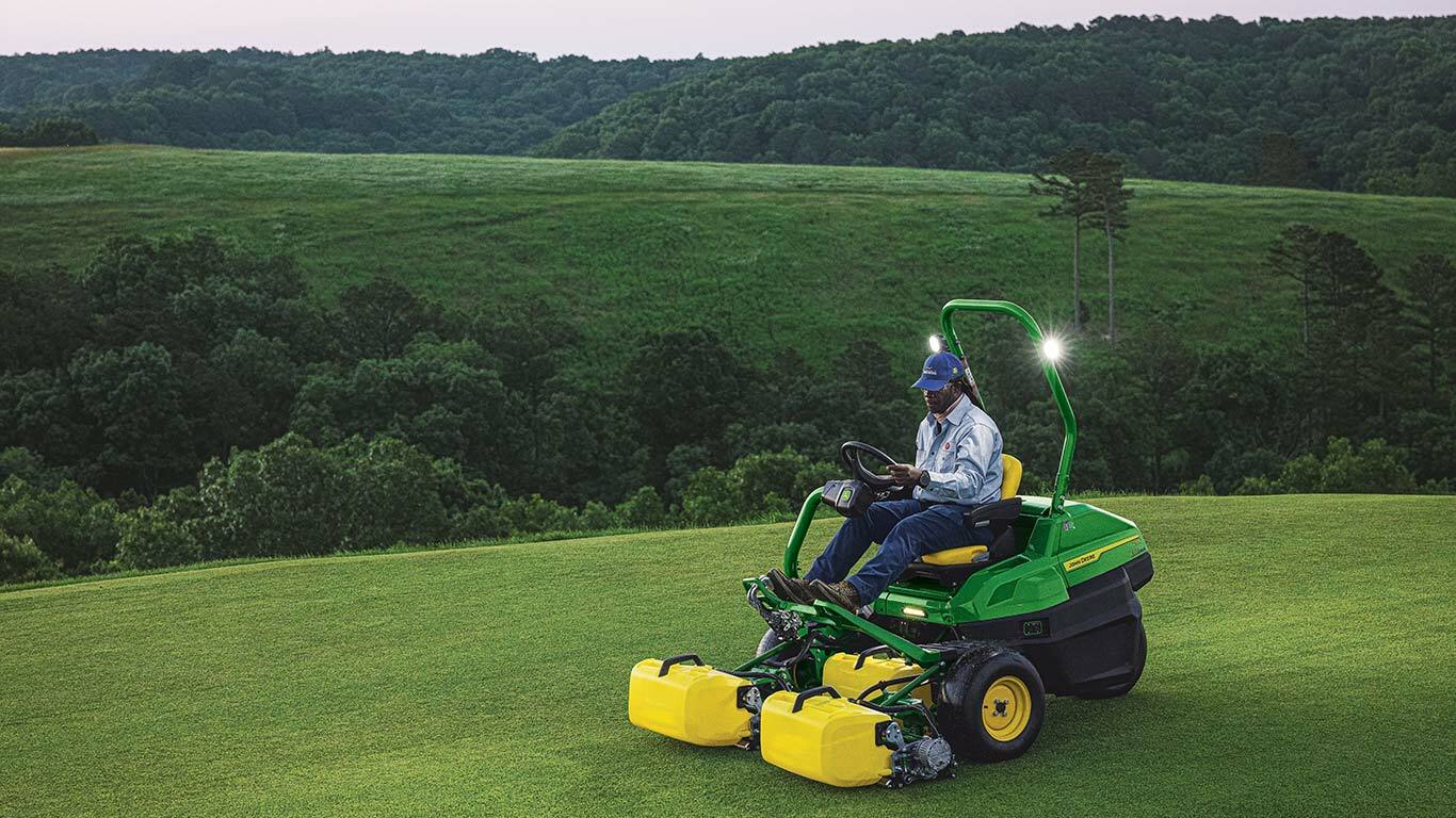 Person mowing golf course with John Deere mower, surrounded by green hills and trees.
