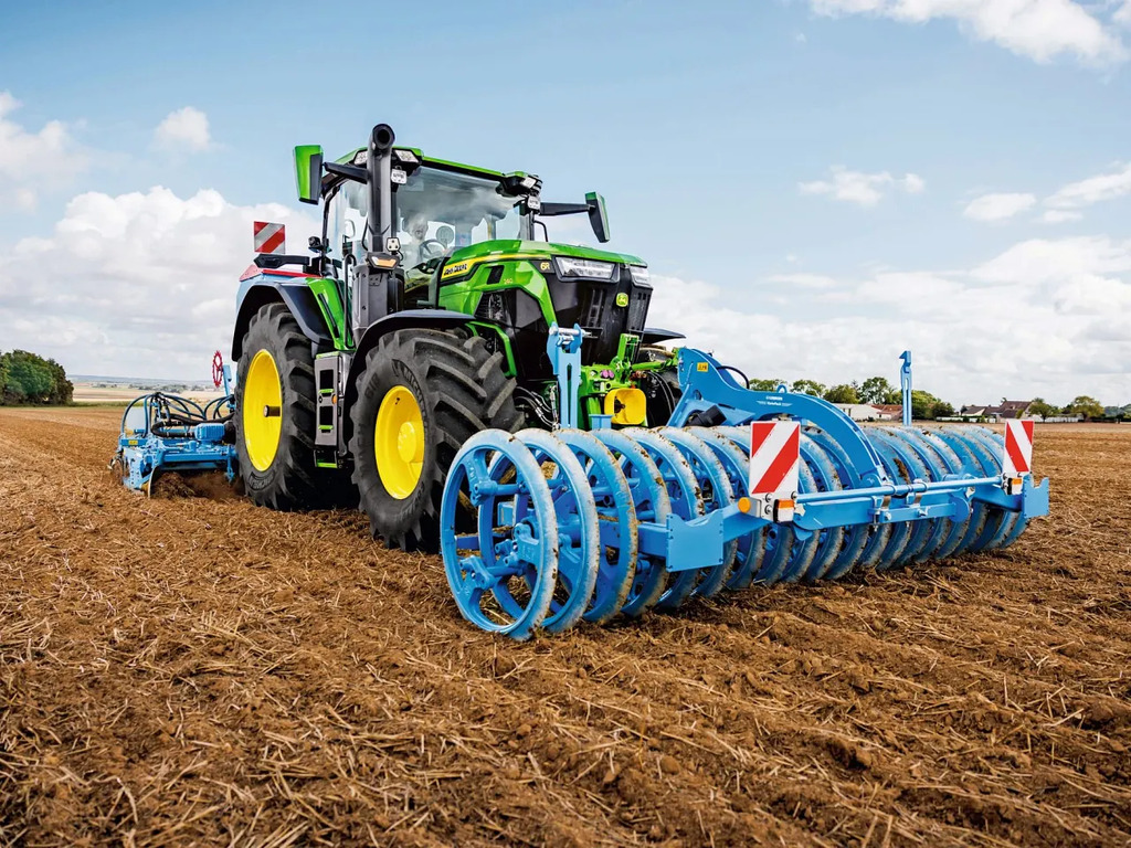 Seedbed preparation with John Deere tractor pulling blue ring roller, firming soil on harvested field under clear sky
