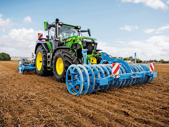 Seedbed preparation with John Deere tractor pulling blue ring roller, firming soil on harvested field under clear sky