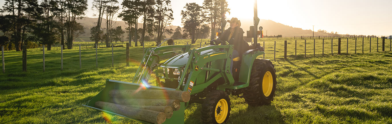 Person drives green tractor with logs in sunny field, trees and hills in background.