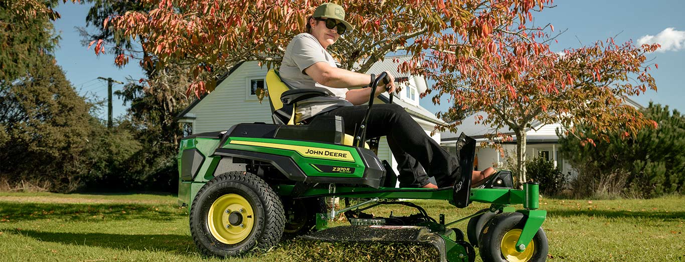 Person rides John Deere Z915E mower on lawn with fall trees and house in background.