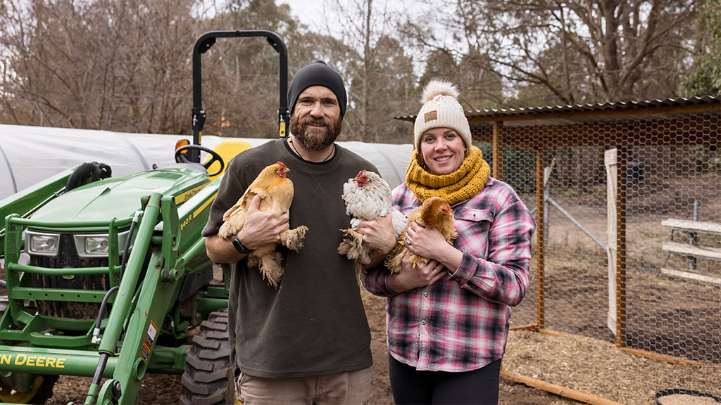 Person in beanie and brown shirt stands with arms crossed in front of green tractor.