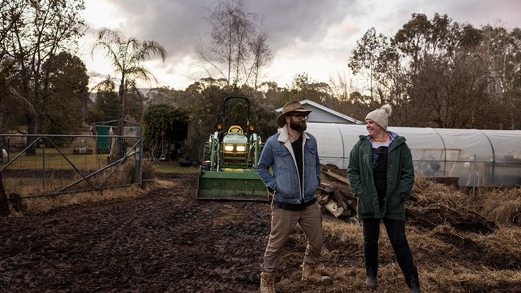Two people in muddy farmyard with tractor and greenhouse in background.
