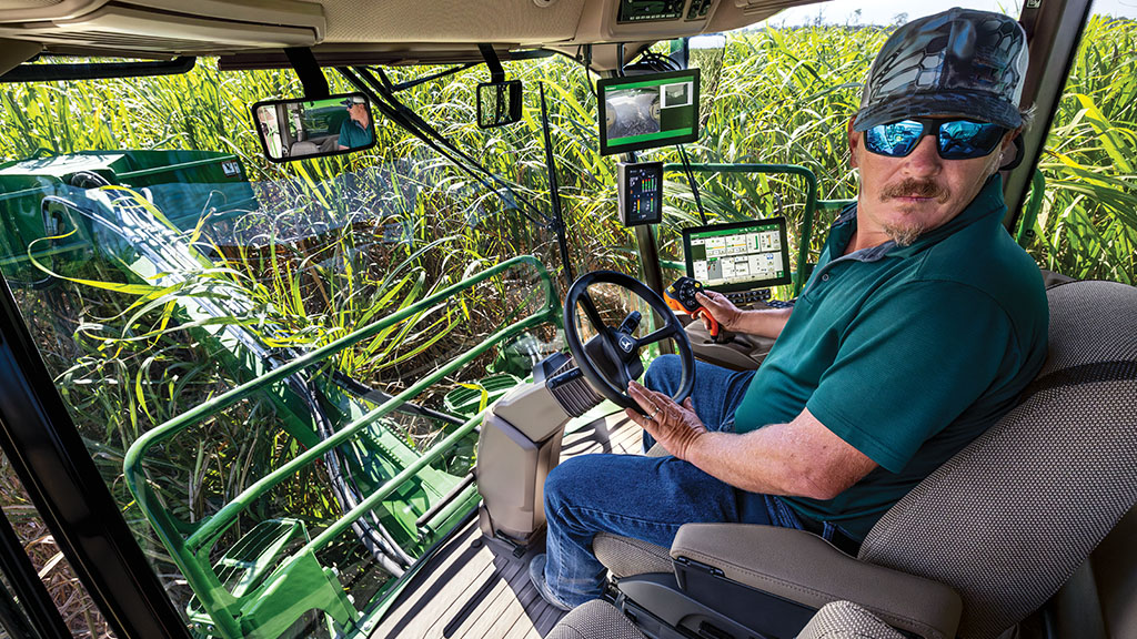 An in-cab, close-up photo showing an operator – in a CH Series Sugar Harvester – turned toward the camera, with standing crops visible through the windows. An in-cab, close-up photo showing an operator – in a CH Series Sugar Harvester – turned toward the camera, with standing crops visible through the windows.
