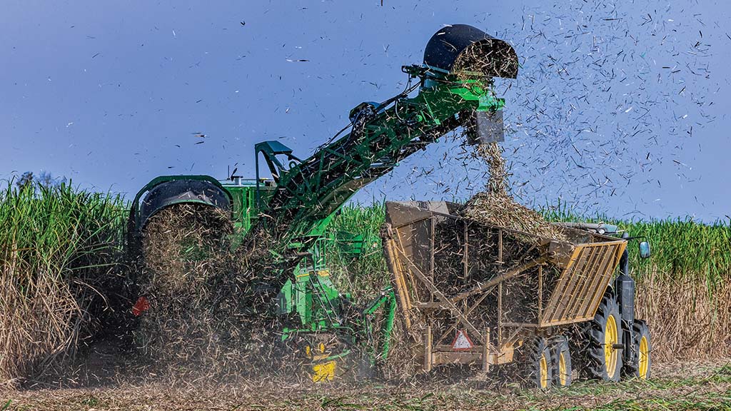 A photo showing CH Series Sugar Harvester unloading sugar cane into a tractor-pulled grain cart, with cane debris floating in the air. A photo showing CH Series Sugar Harvester unloading sugar cane into a tractor-pulled grain cart, with cane debris floating in the air.