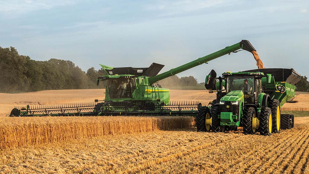 Combine harvesting crop, automatically nudging John Deere tractor to move the grain cart forward and backward to evenly fill.