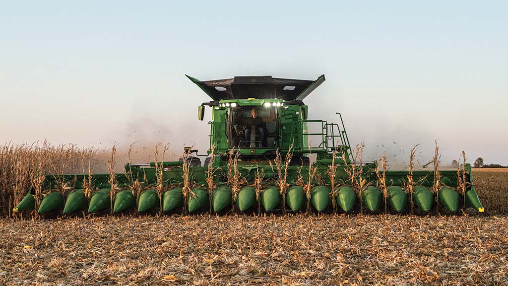 Combine harvester harvesting corn in a field, with corn stalks visible at the front, under a clear sky at dusk. Combine harvester harvesting corn in a field, with corn stalks visible at the front, under a clear sky at dusk.