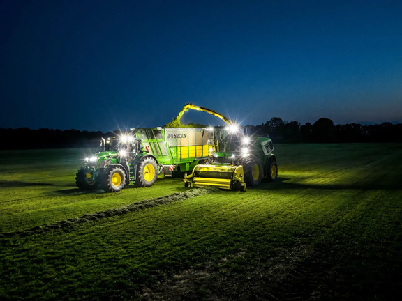 Night grass silage harvesting with John Deere tractor, forage harvester and silage wagon working under field lights