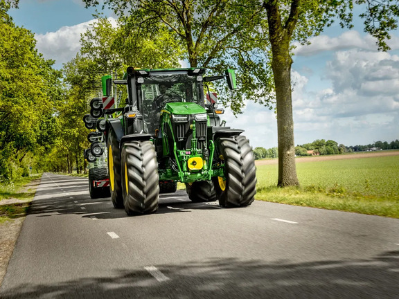 John Deere tractor towing farm equipment on a rural country road beside green fields and trees