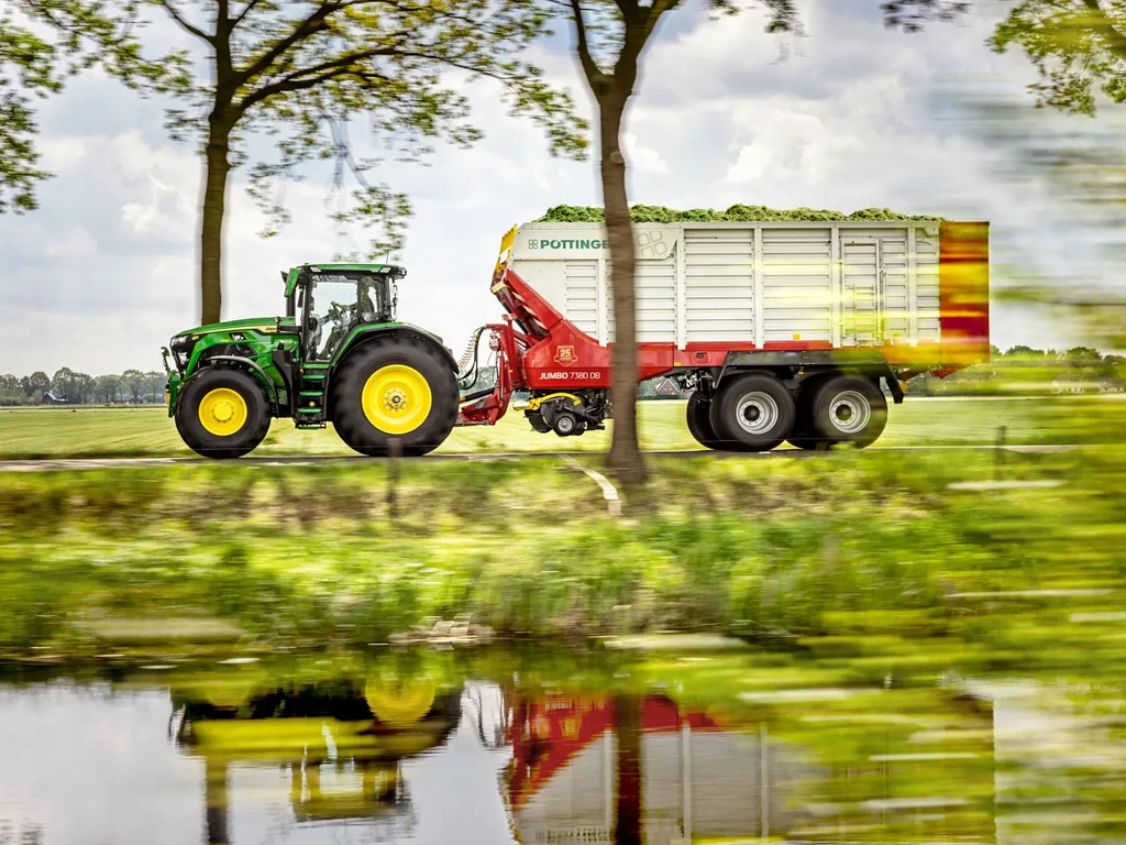 Forage transport with John Deere tractor and Pöttinger silage wagon driving along a rural road by a canal