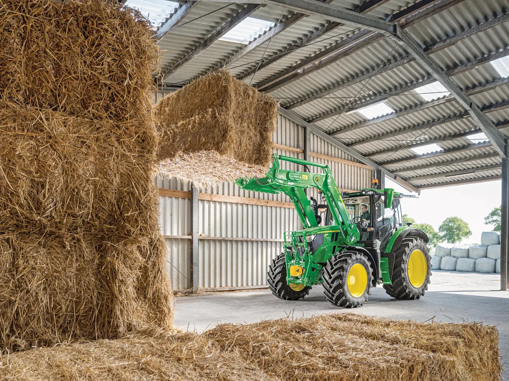 John Deere 6R tractor using a front loader to lift a large straw bale inside a farm shed, stacking hay for storage.