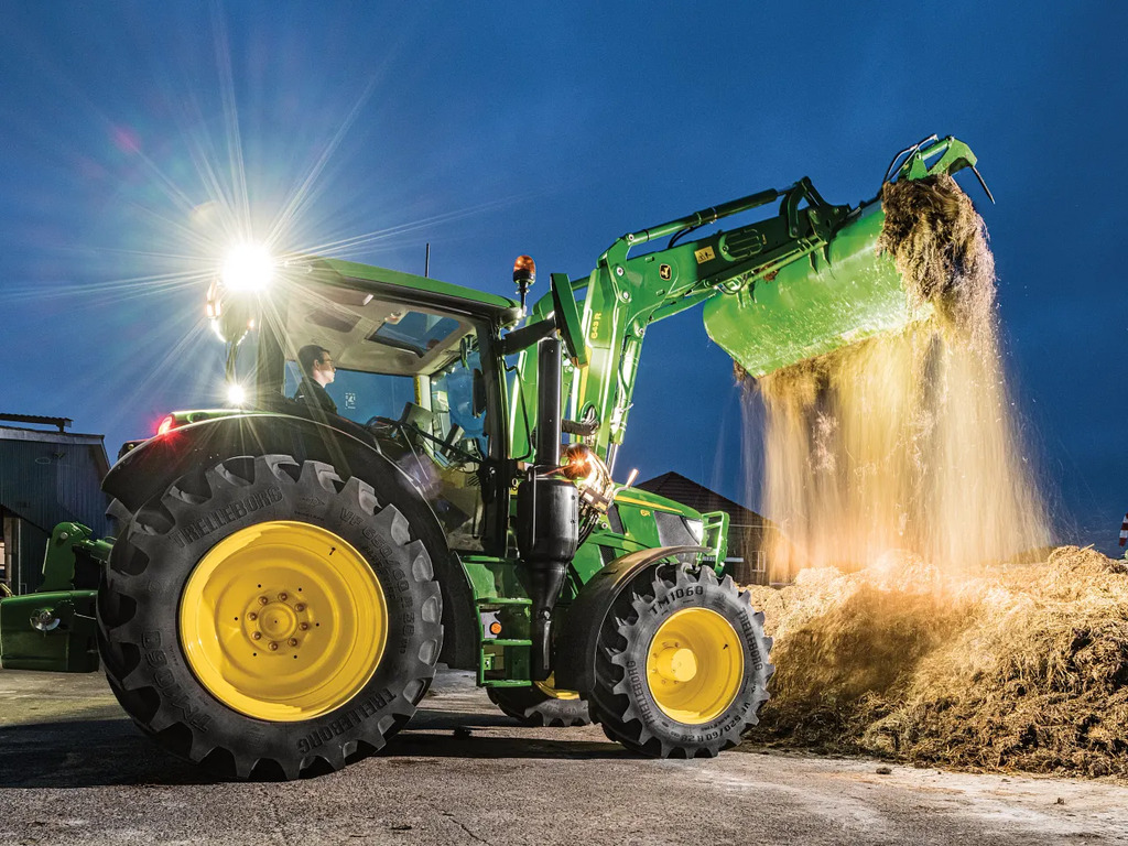 John Deere 6R tractor using a front loader to lift and dump straw or feed at dusk, with bright work lights illuminating the farmyard.