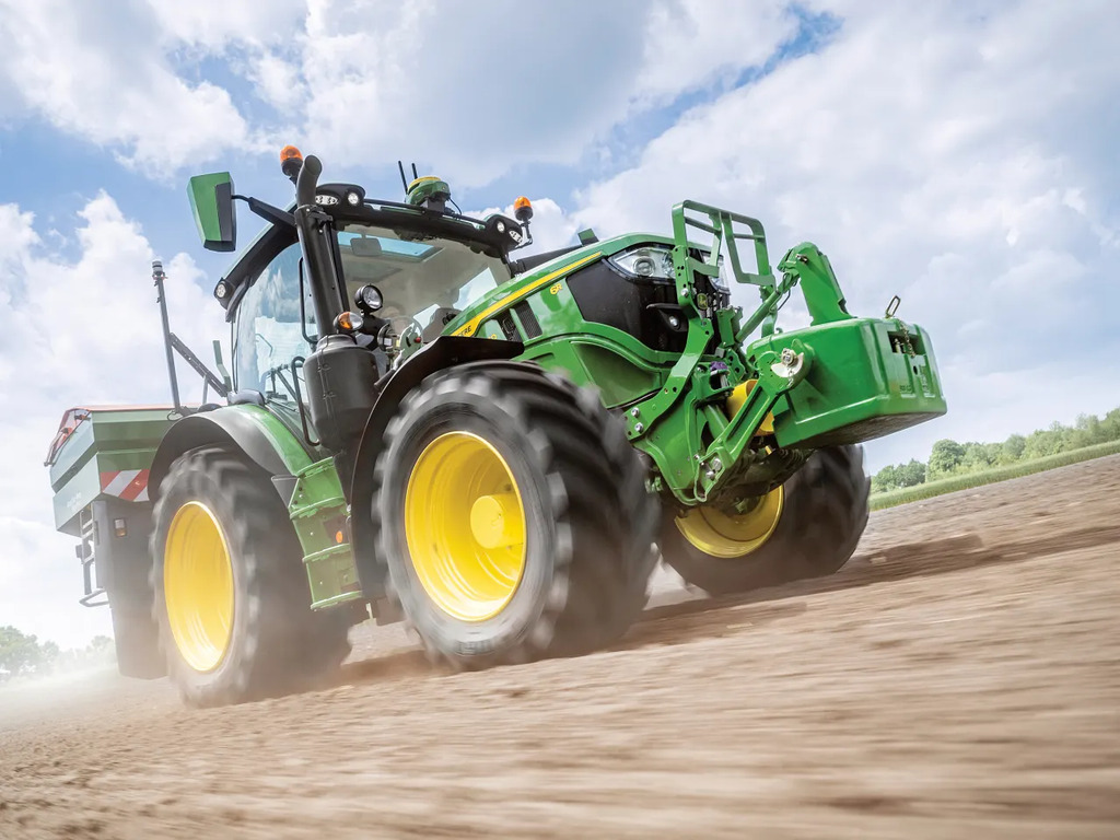 John Deere 6R tractor driving across a dry soil field with a front weight block, creating dust while preparing land for work
