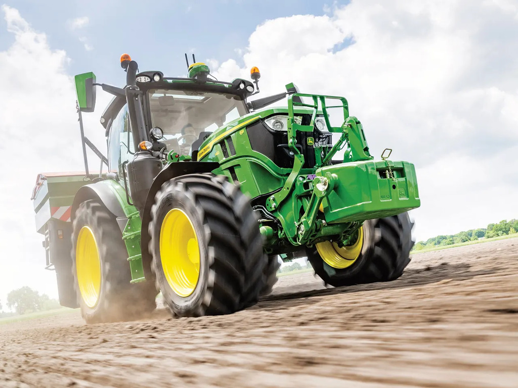John Deere 6R tractor accelerating across a dry soil field with a front weight block, kicking up dust during land preparation.