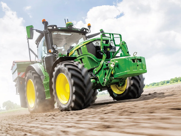 John Deere 6R tractor accelerating across a dry soil field with a front weight block, kicking up dust during land preparation.