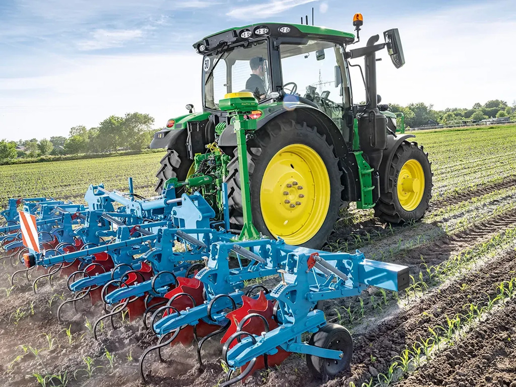 John Deere tractor cultivating young crop rows with a rear‑mounted inter-row cultivator on a sunny field.