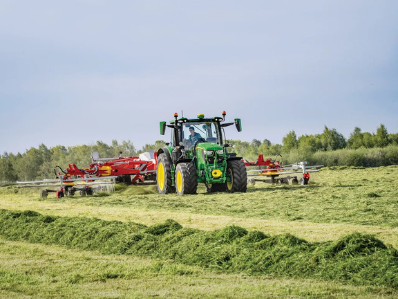 John Deere 6R tractor working in a field with a large rake attachment, spreading cut grass during haymaking on open farmland.