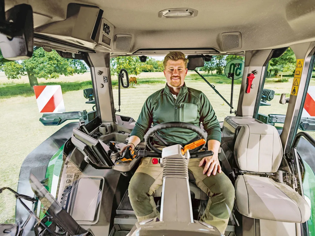 Tractor operator seated inside a John Deere cab, hands on steering wheel and controls, with full interior visibility and safety markings